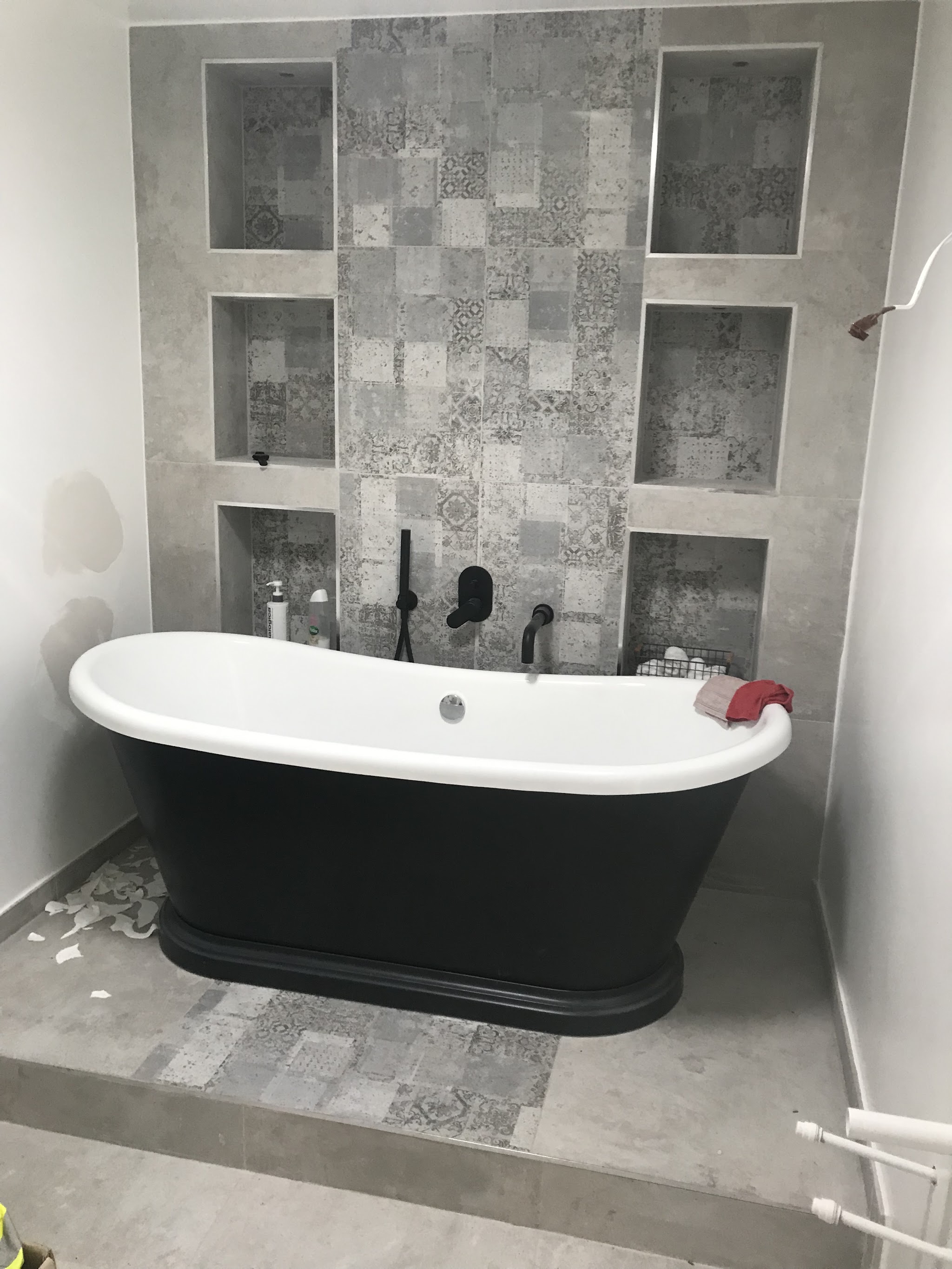 Black freestanding slipper bath on a raised patterned-tile plinth with tiled niches behind